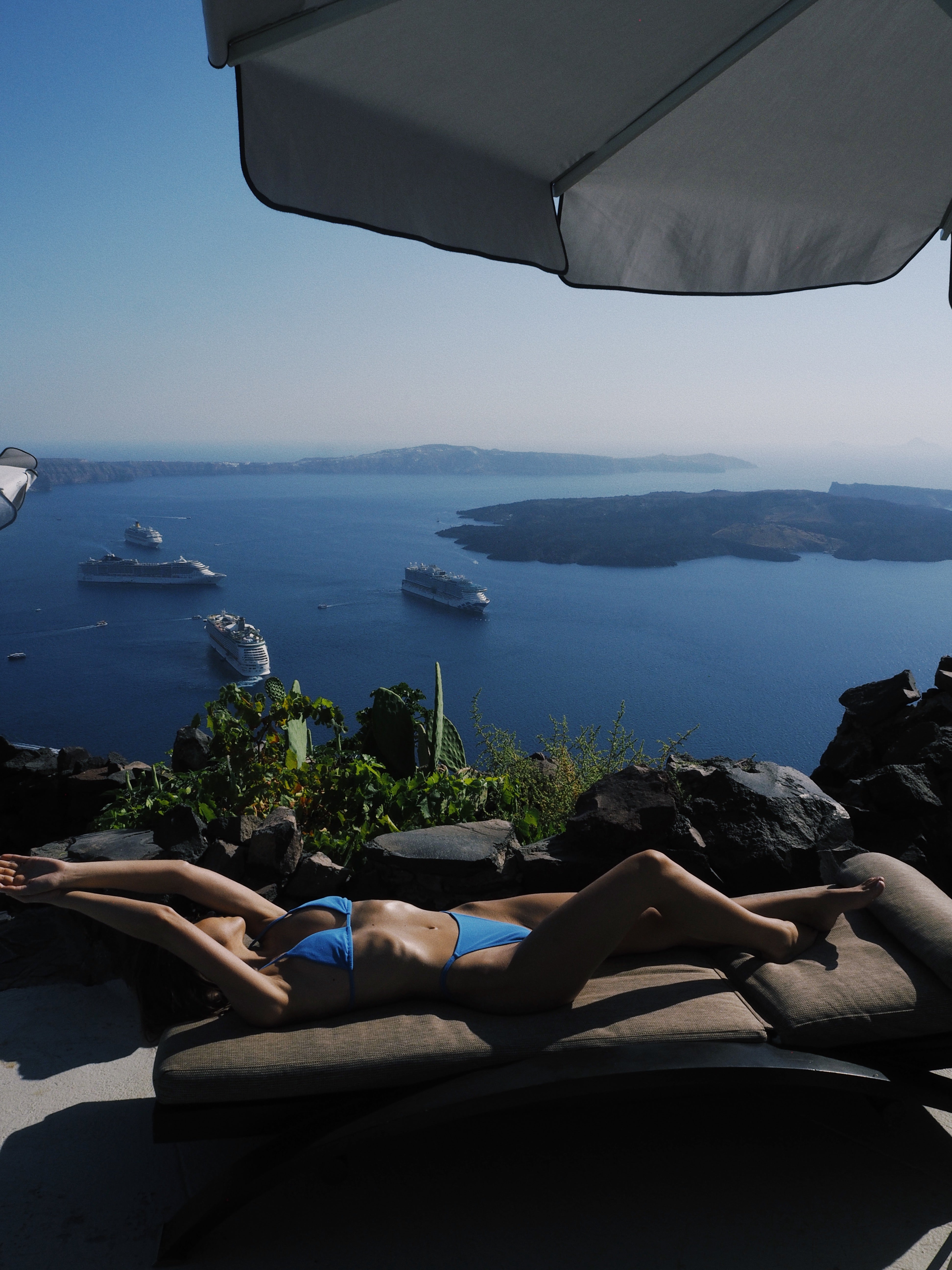 Person in a blue bikini lying on a sun lounger with a scenic view of the ocean and islands.